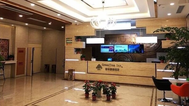A modern hotel reception area with a wooden reception desk, a digital screen, and potted plants.