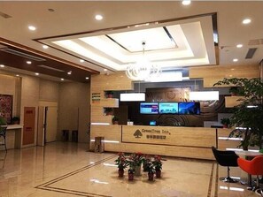 Una moderna zona de recepción de hotel con un mostrador de madera, una pantalla digital y plantas en macetas.