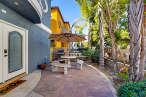 Un espace patio avec des sièges d’extérieur et un barbecue, entouré de palmiers et d’un bâtiment bleu.