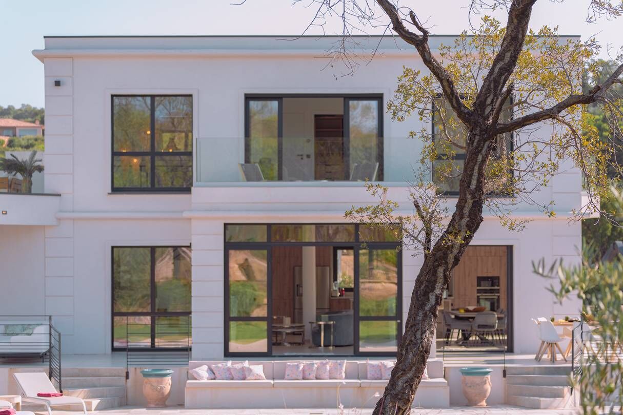 A modern two-story house with large windows, a balcony, and outdoor seating.