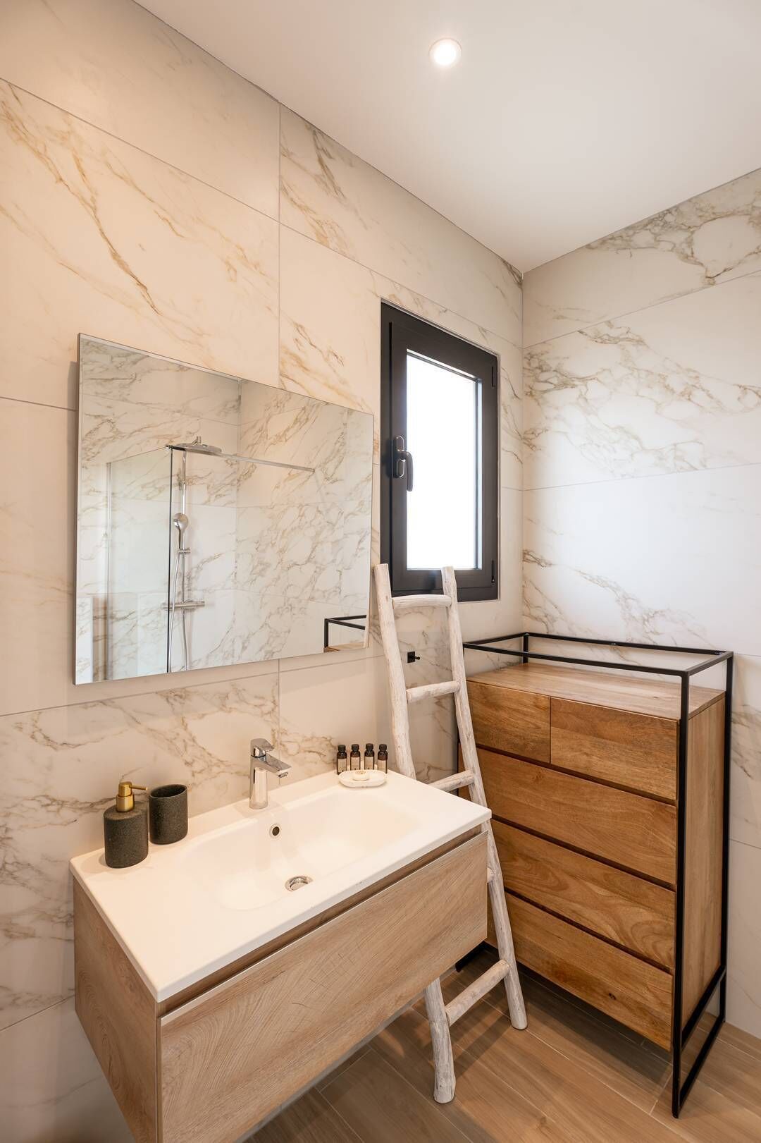 A modern bathroom with a marble wall, a large mirror, a wooden vanity, and a ladder.