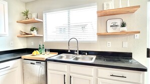 A kitchen with white cabinets, a black countertop, and a window with blinds. There are wooden shelves above the counter with decorative items. - Contemporary 4-Bed Home Steps from Downtown & ATT Center (San Antonio)