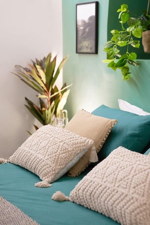 Une chambre à coucher bien rangée, avec une literie de couleur turquoise, des coussins blancs à texture et une plante suspendue.