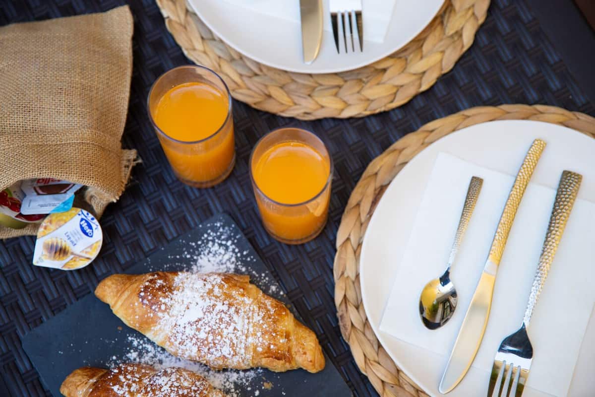 Un desayuno con un croissant, dos vasos de jugo de naranja, un plato con cubiertos y un recipiente pequeño de yogur.
