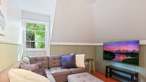 Ein Wohnzimmer mit einem großen Flachbildfernseher, einem grauen Sofa mit vielen Kissen, einer Holzbank und einem Fenster mit Blick auf Grünflächen.
