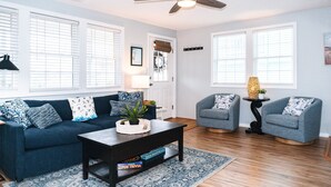 A living room with a dark blue sofa, two grey armchairs, a coffee table, and a ceiling fan. - Classic Beach Cottage with front porch view of water (Carolina Beach)
