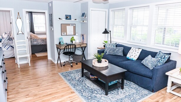 A well-lit living room with a blue sofa, a coffee table, and a ceiling fan. There is a desk with a chair and a bookshelf in the background. - Classic Beach Cottage with front porch view of water (Carolina Beach)