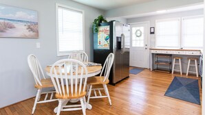 A kitchen with a dining table, chairs, a refrigerator, and a window with blinds. - Classic Beach Cottage with front porch view of water (Carolina Beach)