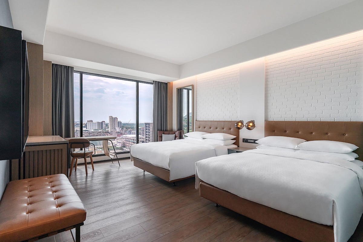 A hotel room with two beds, a desk, a chair, and a view of the city.