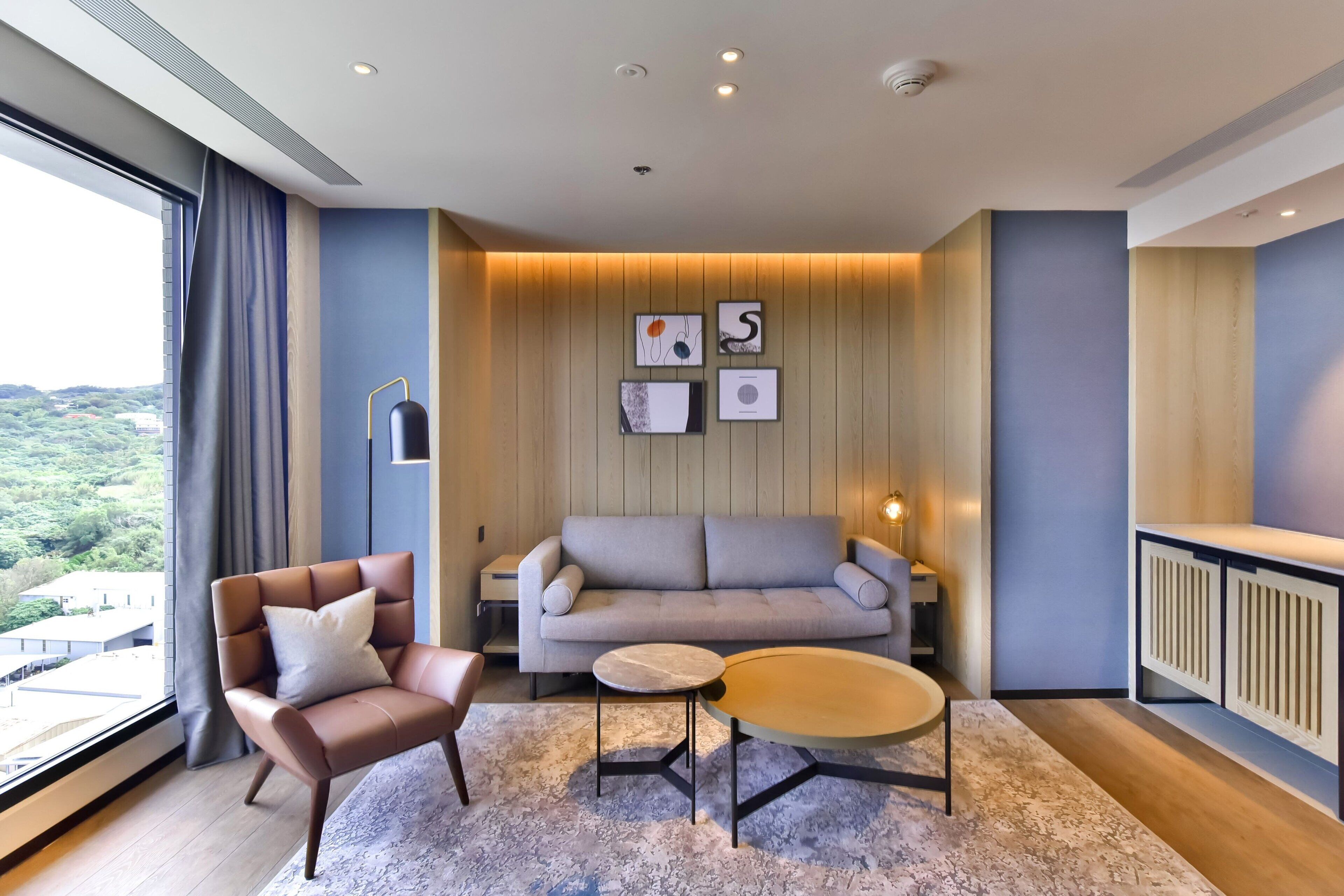 A modern living room with a sofa, armchair, and coffee table, a large window with curtains, and a city view.