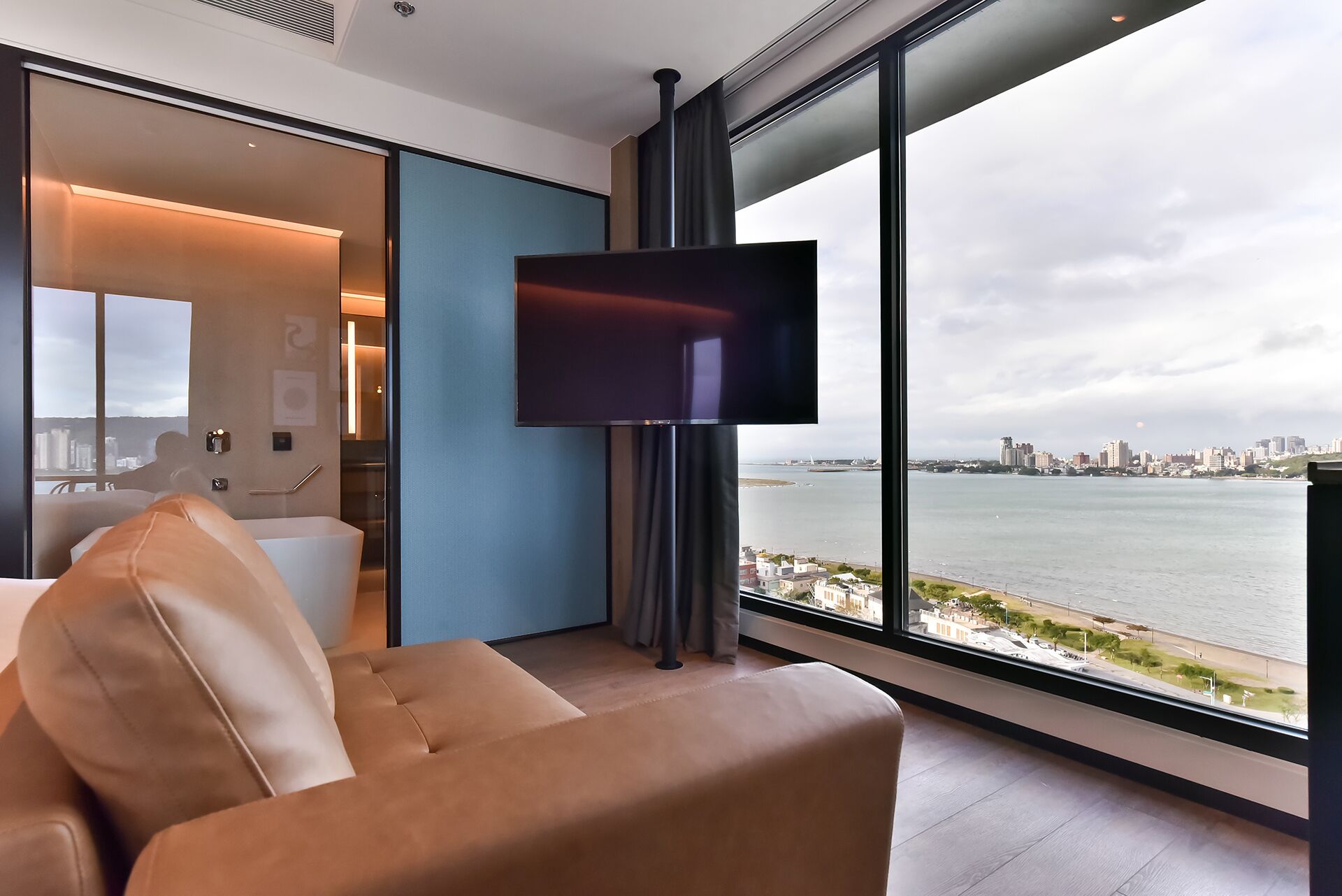 A modern hotel room with a large window offering a city view, a flat-screen TV mounted on the wall, and a comfortable seating area with a sofa and a chair.
