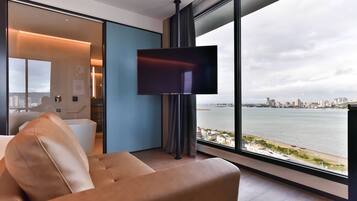 A modern hotel room with a large window offering a city view, a flat-screen TV mounted on the wall, and a comfortable seating area with a sofa and a chair.