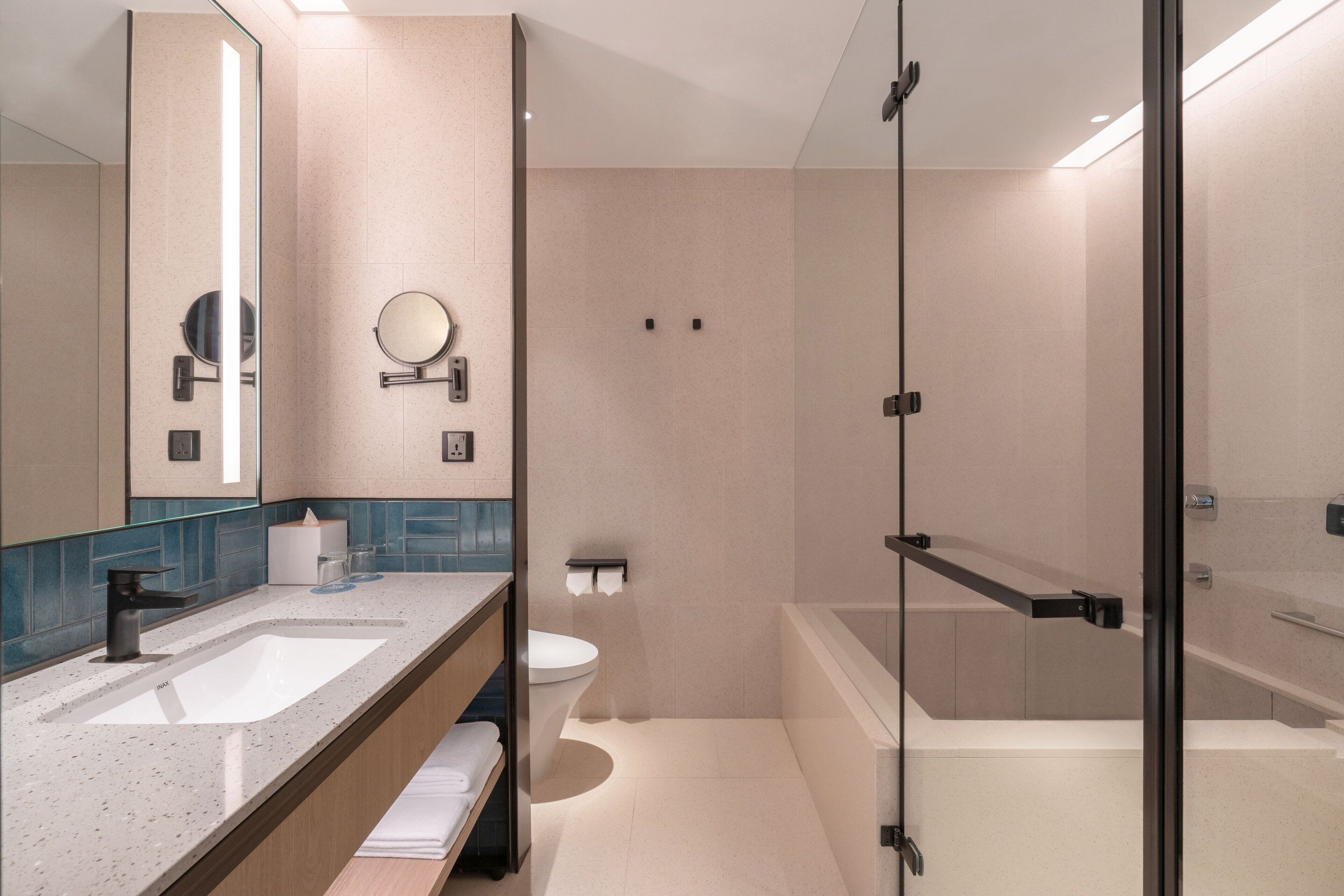 A modern bathroom with a large mirror, two sinks, a toilet, and a glass-enclosed shower area.