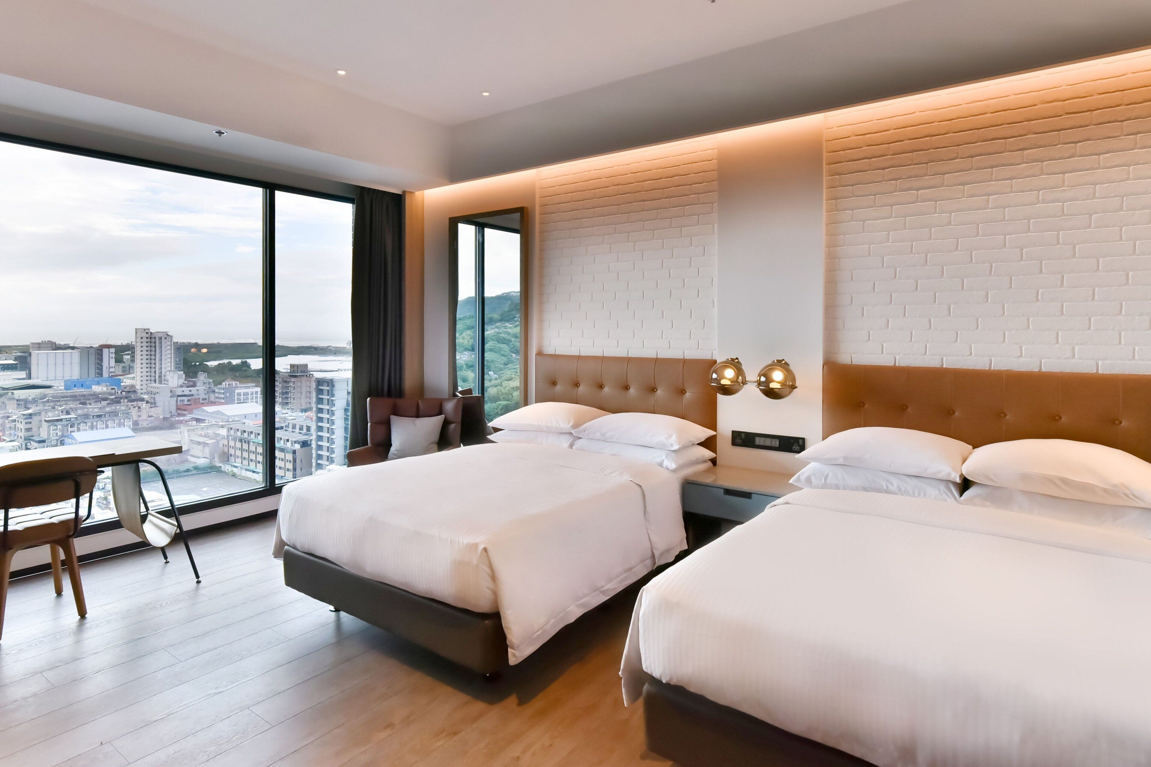 A hotel room with two beds, a desk, and a large window offering a city view.