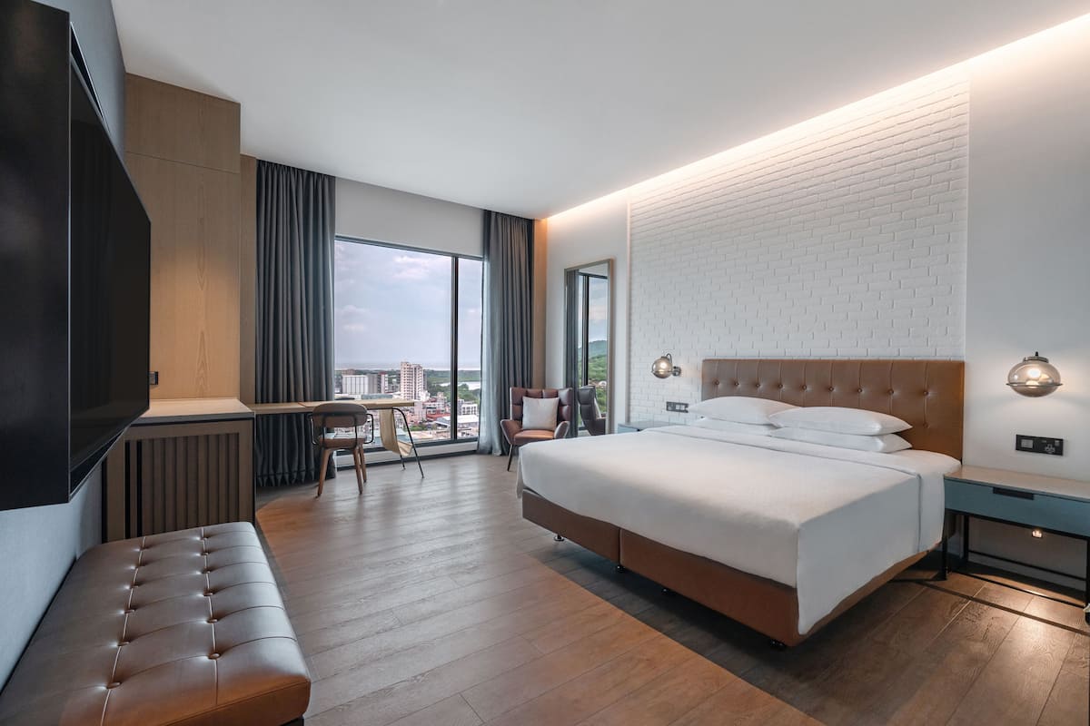 A modern hotel room with a large bed, a seating area, a desk, and a view of the city.
