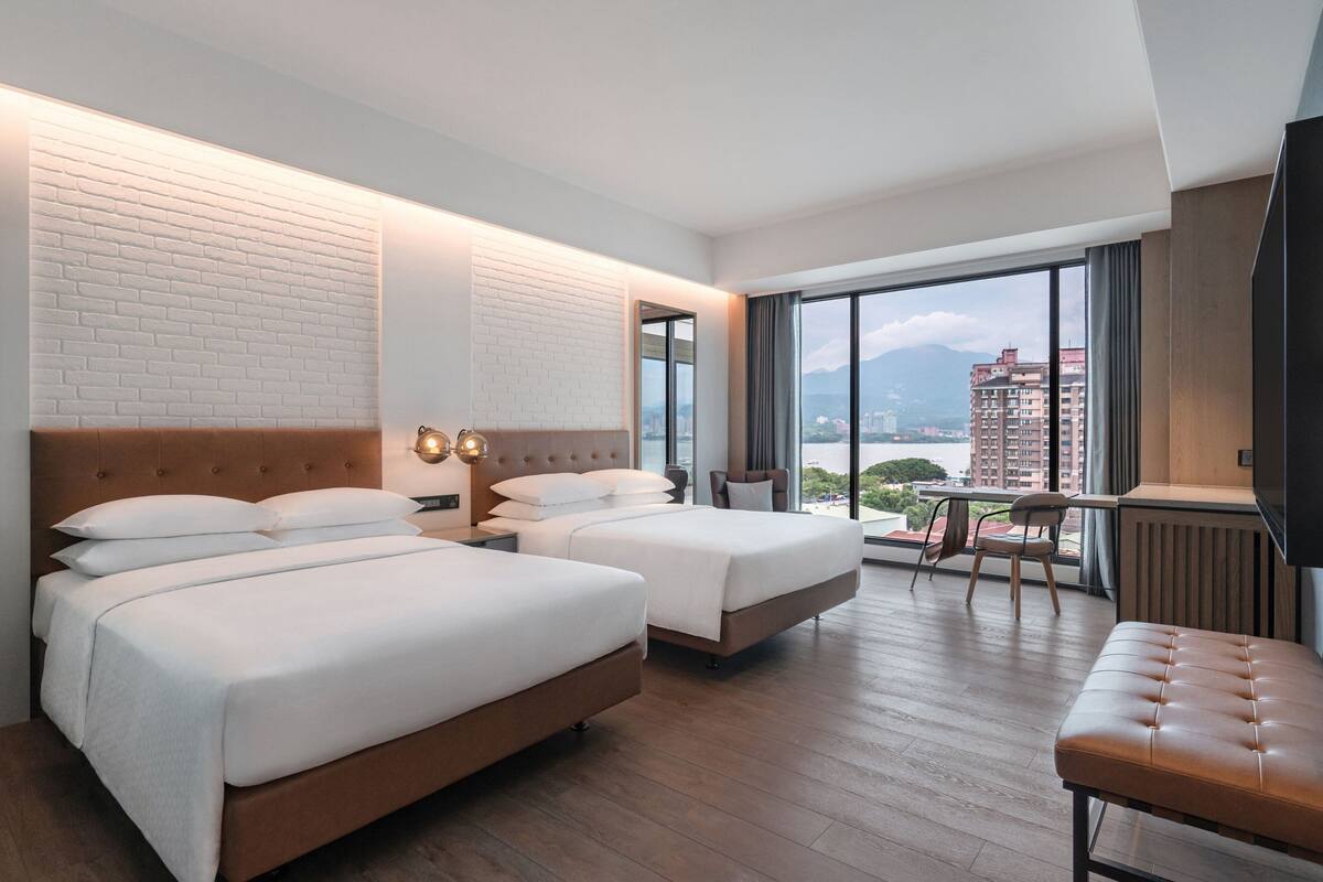 A hotel room with two beds, a wooden bench, a desk, and a large window offering a city view.