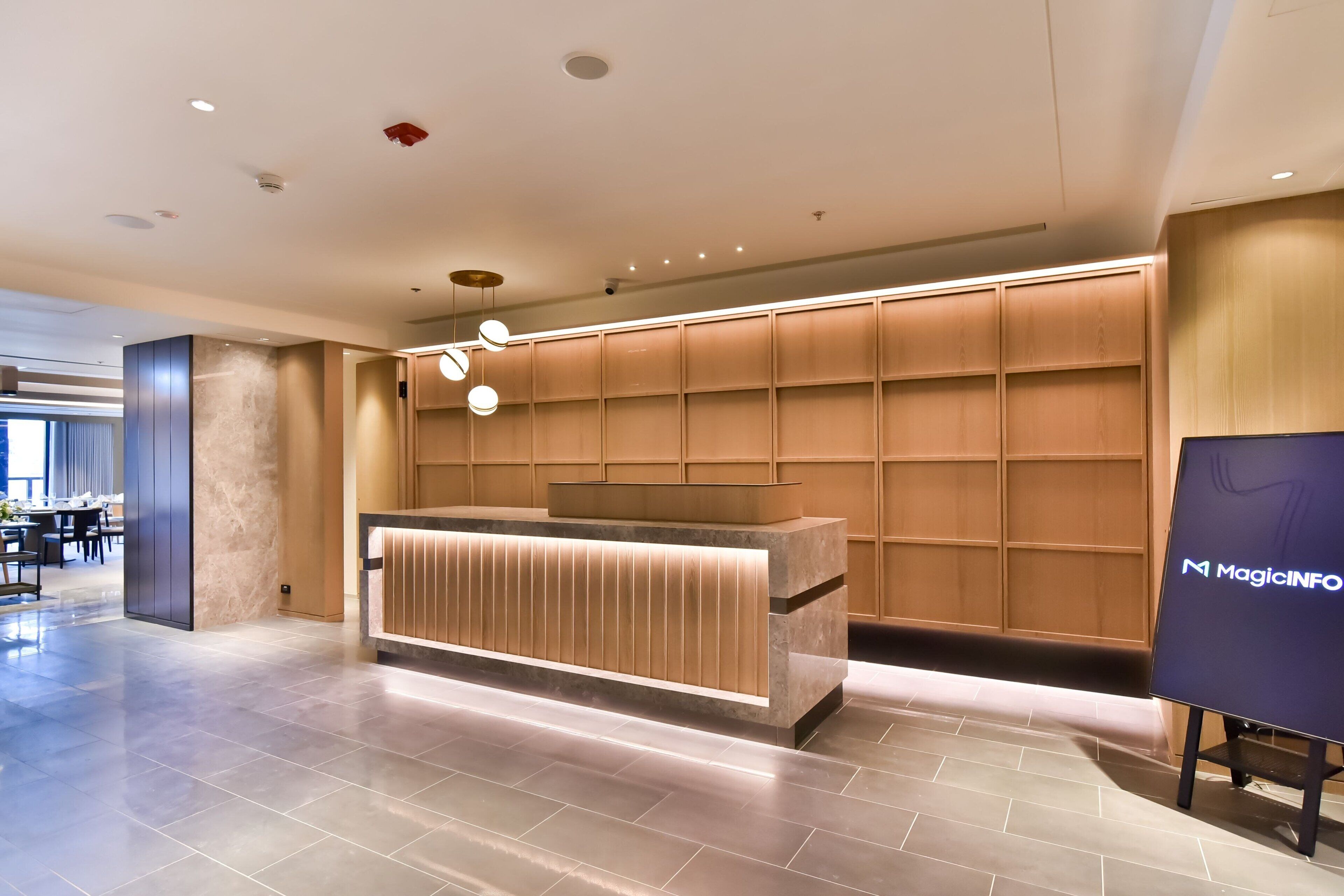 A modern reception area with a wooden reception desk, a wall-mounted information board, and a tiled floor.