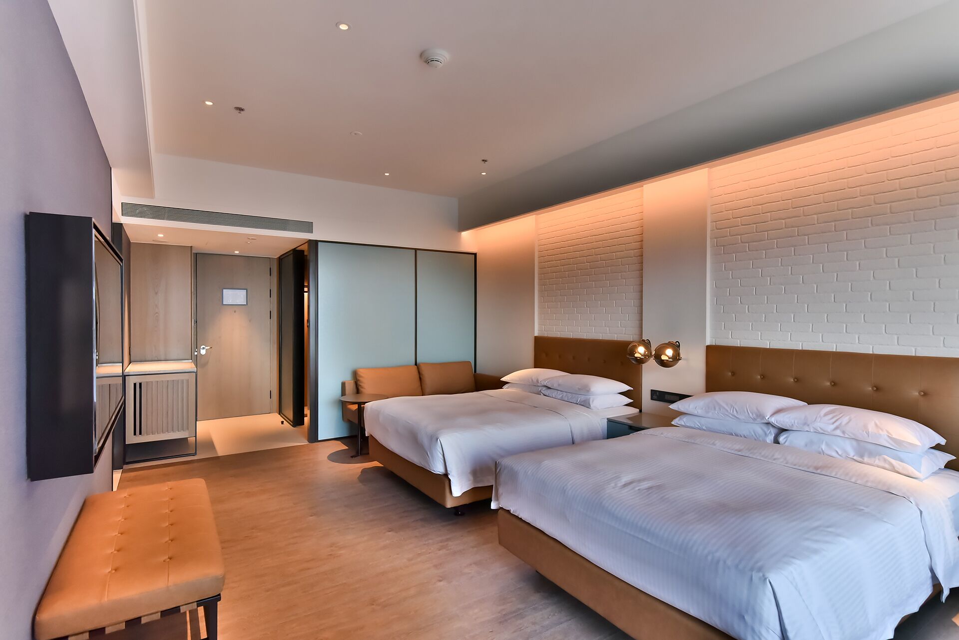 A modern hotel room with two beds, a bench, a wardrobe, and a large mirror.