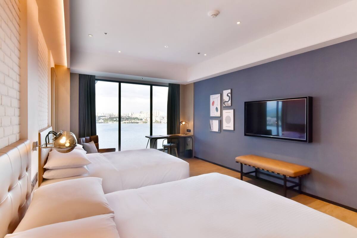 A hotel room with two beds, a desk, a bench, a TV, and a large window with a city view.