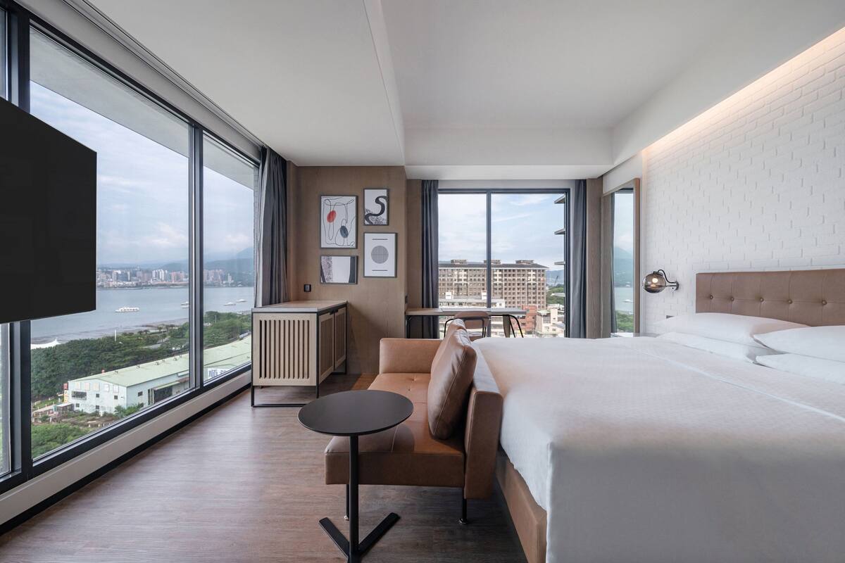 A modern hotel room with a large bed, a sofa, a small table, and a view of the city and water.