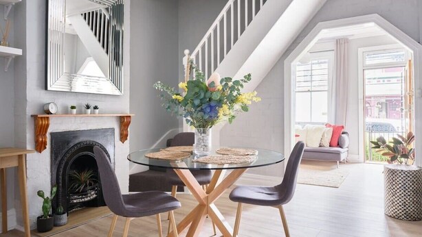 Un comedor con mesa de vidrio, cuatro sillas, un jarrón con flores, chimenea y escalera.
