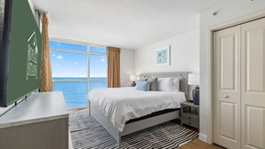 Quarto com cama, mesa de cabeceira, abajur e vista para o mar.