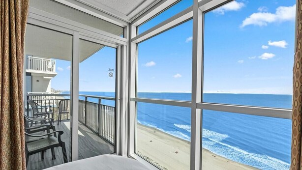 Quarto com grande janela com vista para a praia, varanda com cadeiras e uma cama.