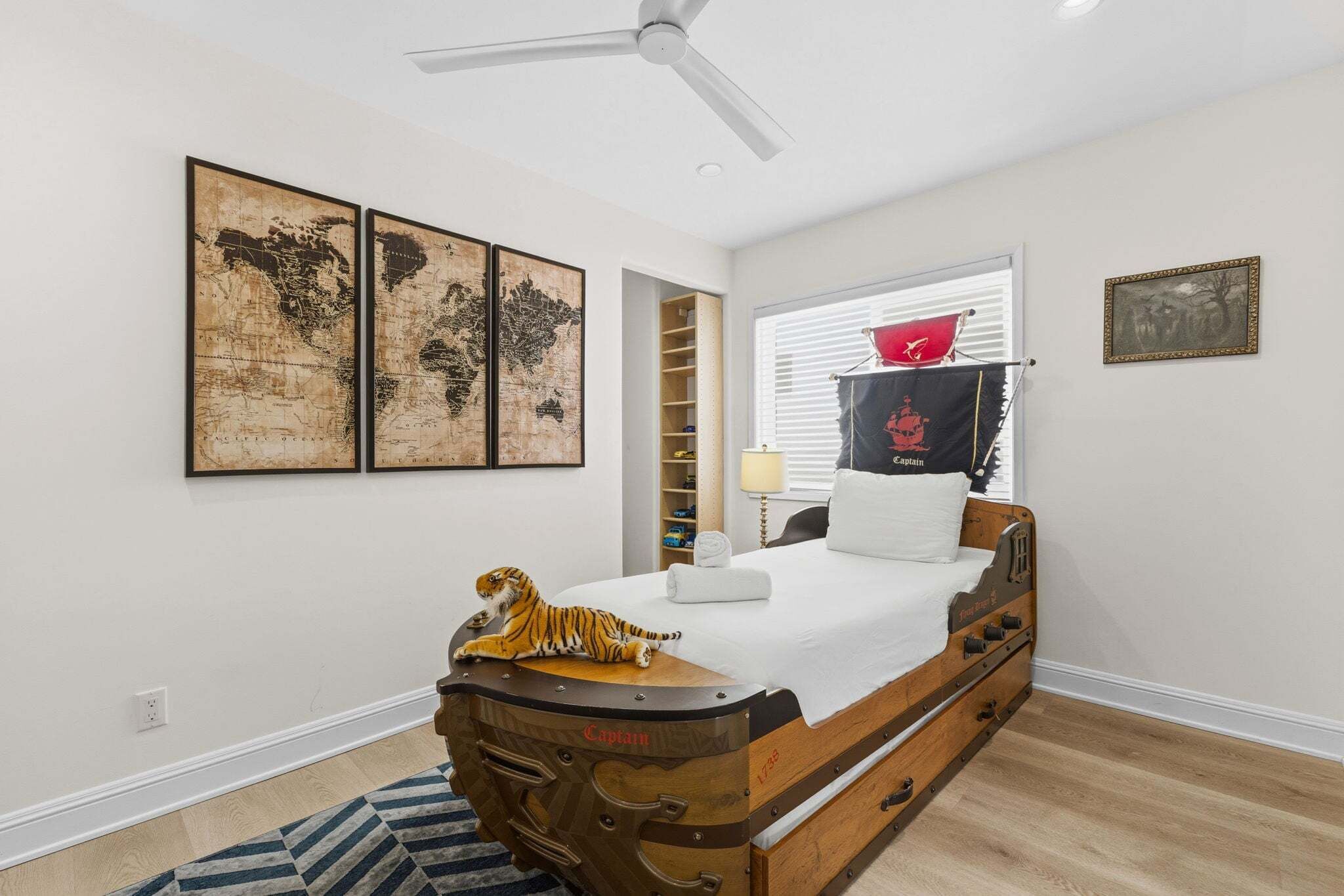 Une chambre avec un lit en bois, un ventilateur de plafond, une carte du monde, un tigre en peluche, un coussin sur le thème du Capitaine, et une fenêtre avec des stores.