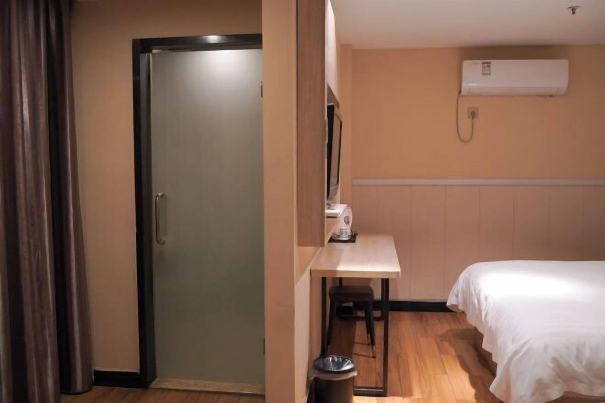 A hotel room with a bed, a desk, a chair, a trash bin, and an air conditioning unit.