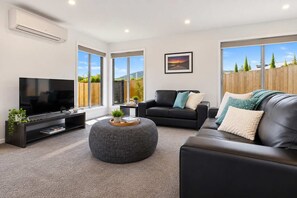 Ein modernes Wohnzimmer mit einem Flachbildfernseher, Ledersofas, einem runden Hocker und großen Fenstern mit Blick ins Freie.