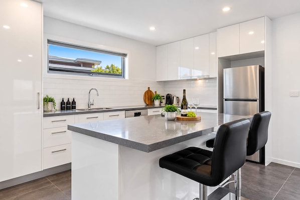 A modern kitchen with white cabinets, a central island with bar stools, a stainless steel refrigerator, and a window with a view of a house.
