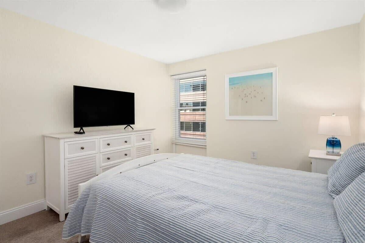 Une chambre à coucher avec un lit, un téléviseur posé sur une commode, une fenêtre avec des stores et un tableau accroché au mur.