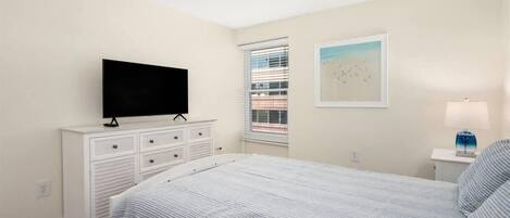 Une chambre à coucher avec un lit, un téléviseur posé sur une commode, une fenêtre avec des stores et un tableau accroché au mur.