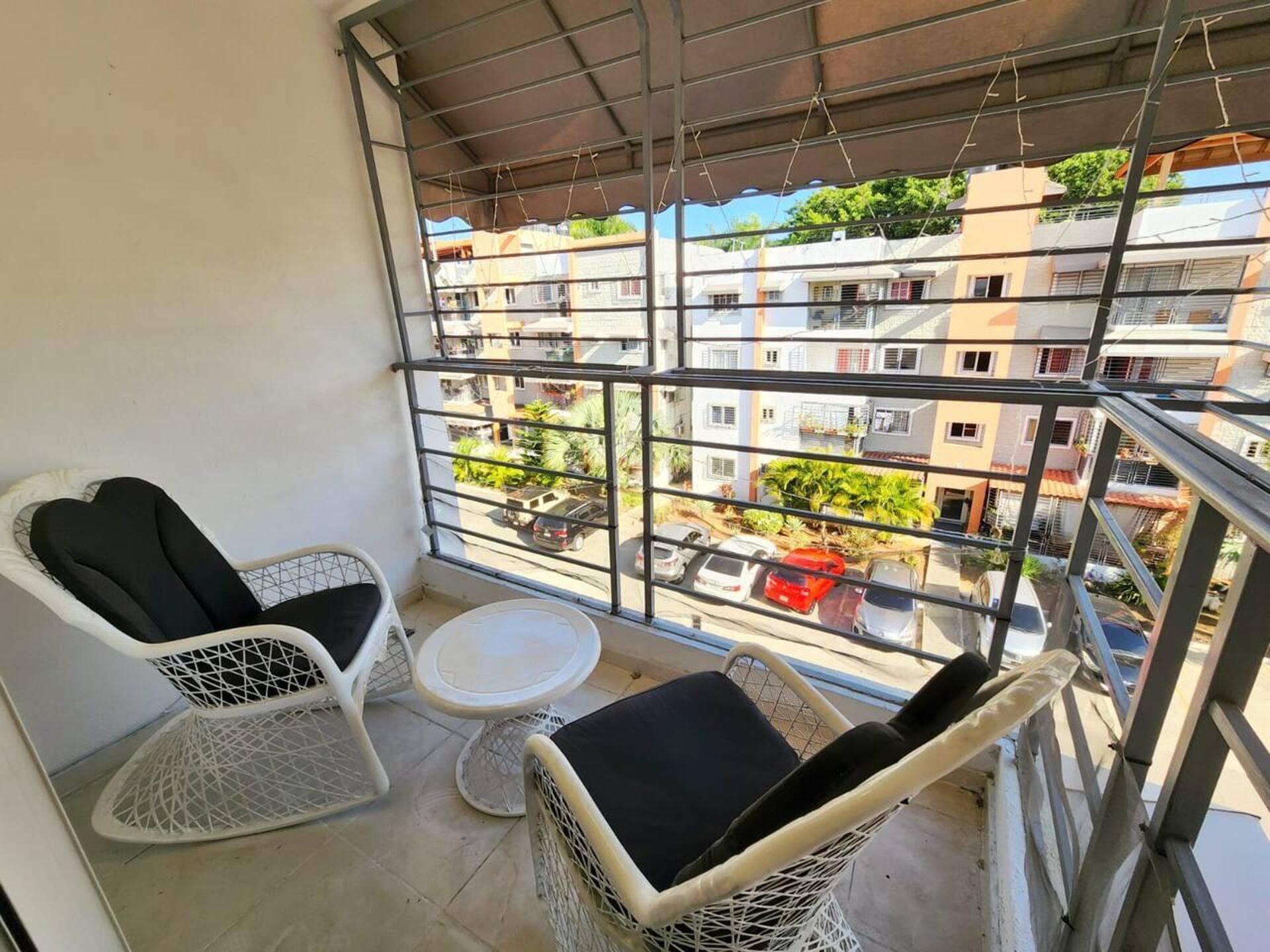 A balcony with two wicker chairs and a small round table, overlooking a residential area with multiple buildings and parked cars. - Charming 3-bed Apartment 2 Bano Aire Acondicionado (Santo Domingo Oeste)