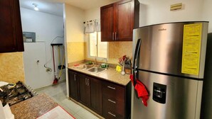 A kitchen with a stainless steel refrigerator, wooden cabinets, a window with a curtain, and a tiled backsplash. - Charming 3-bed Apartment 2 Bano Aire Acondicionado (Santo Domingo Oeste)