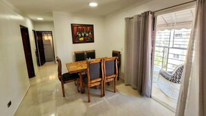 A dining area with a wooden table and chairs, a painting on the wall, and a view of a balcony with a white chair. - Charming 3-bed Apartment 2 Bano Aire Acondicionado (Santo Domingo Oeste)