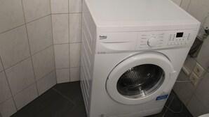 A front-loading white washing machine with a control panel, located in a corner of a room with tiled walls and floor. - Stay2Night Hostel (Zürich)