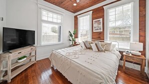 Une chambre avec un plafond en bois, un grand lit avec une literie blanche, un téléviseur posé sur un meuble en bois et des fenêtres équipées de stores.