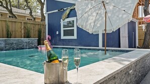 Une piscine avec un seau en métal contenant une bouteille de champagne et deux verres, un parasol blanc et une clôture en bois en arrière-plan.