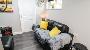 A cozy living room with a black leather sofa, two yellow cushions, a small black stool, a fireplace, and a wall clock. - Lockdown Lodge (Mablethorpe)