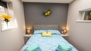 A neatly made bed with a 'Happy' pillow, two folded towels, and a wall-mounted flower wreath. - Lockdown Lodge (Mablethorpe)