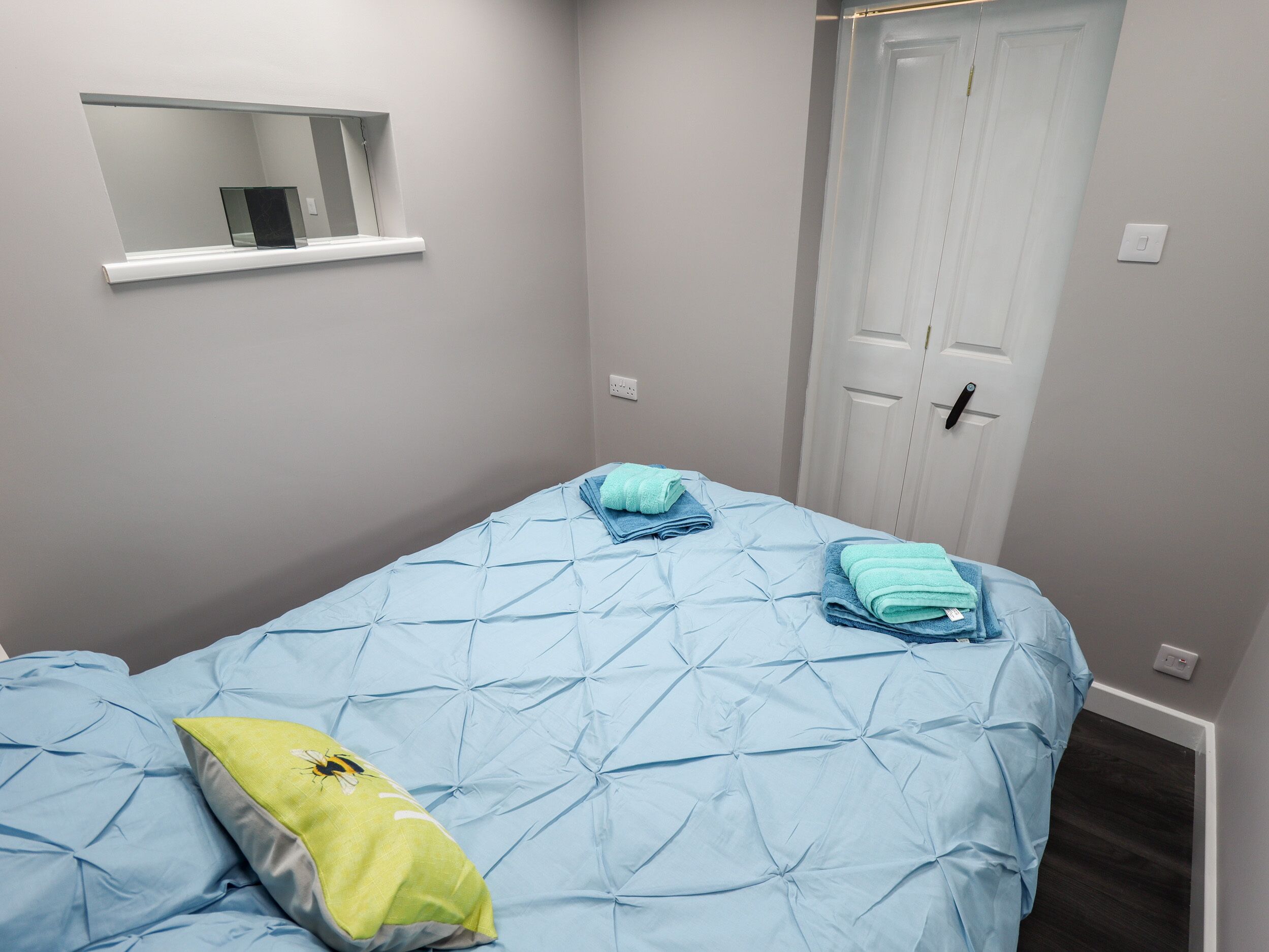 A bedroom with a single bed, a window with a mirror, a door, and a light switch on the wall.