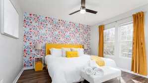 A bedroom with a floral wallpaper, a yellow headboard, a white bed with rolled towels, a bedside table with a lamp, and a window with curtains. - Candy Land- Downtown ILM (Wilmington)