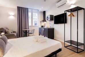 Ein modernes Hotelzimmer mit Bett, einer Couch, einem kleinen Esstisch, einem Fernseher und einer Küchenzeile.
