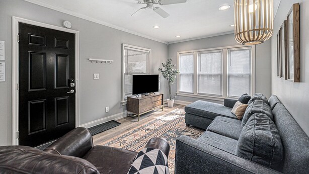 A living room with a black door, a ceiling fan, a TV on a wooden stand, a grey sofa, and a patterned rug. - Updated Modern & Spacious 2 bed Apartment Near Everything! (Jackson)