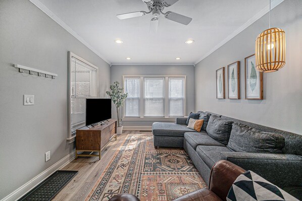 Un salon avec un canapé gris, un téléviseur, un ventilateur de plafond et un tapis à motifs.
