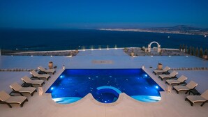 Une piscine à débordement située sur le toit, avec des chaises longues, des plantes en pot et une vue sur la côte au crépuscule.
