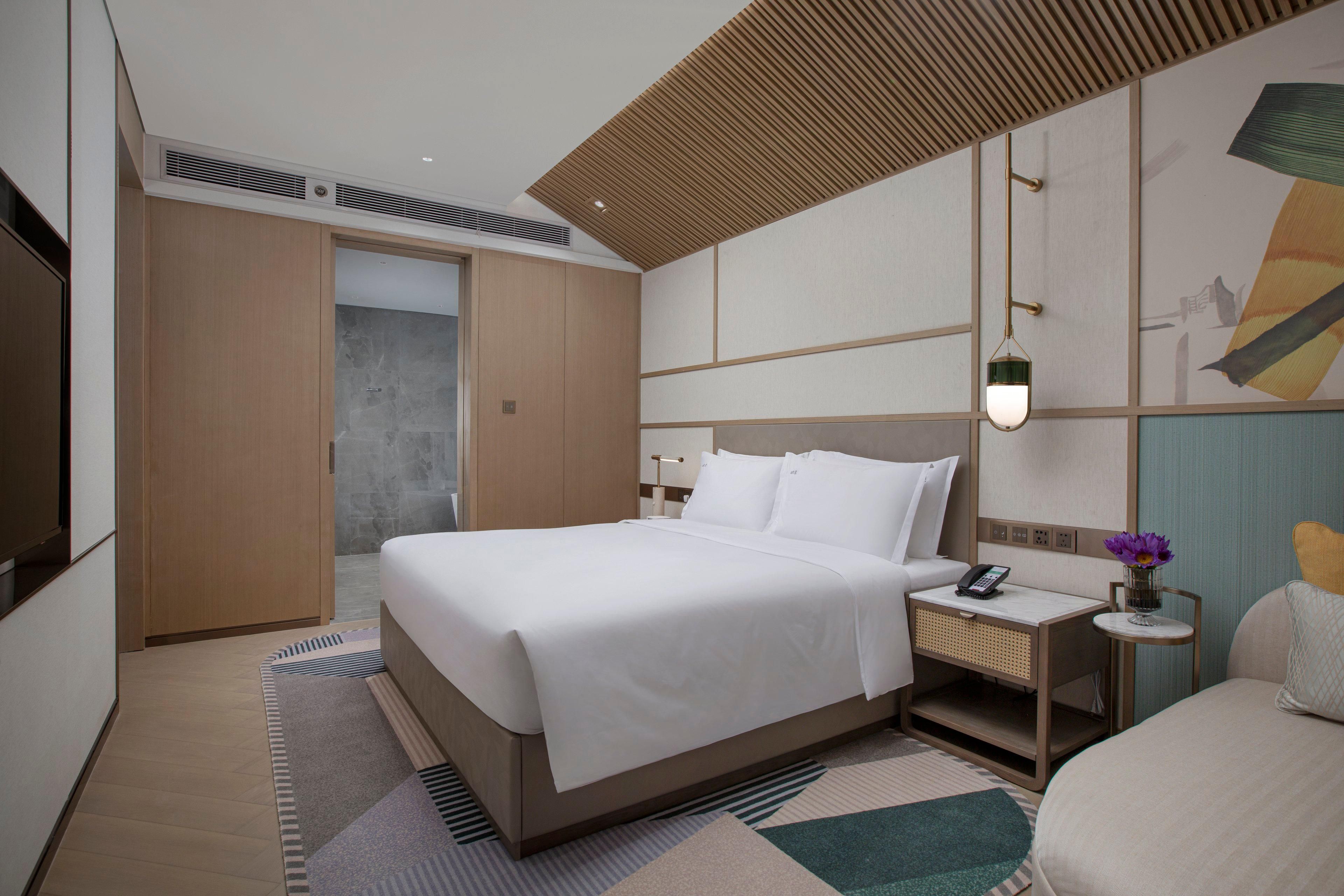 A modern hotel room with a large bed, bedside table, and a small bedside table with a lamp.