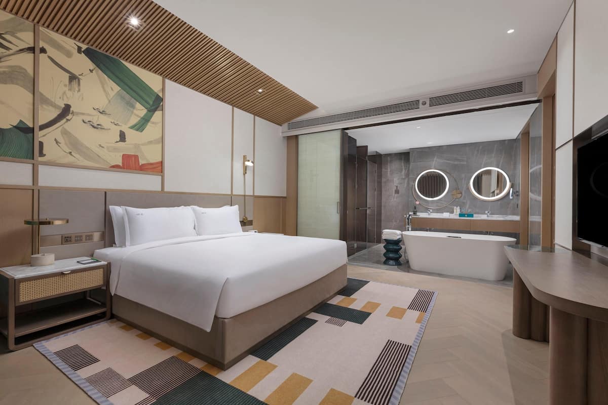 Una habitación de hotel moderna con una cama grande, una mesita de noche y un baño con bañera y un espejo amplio.