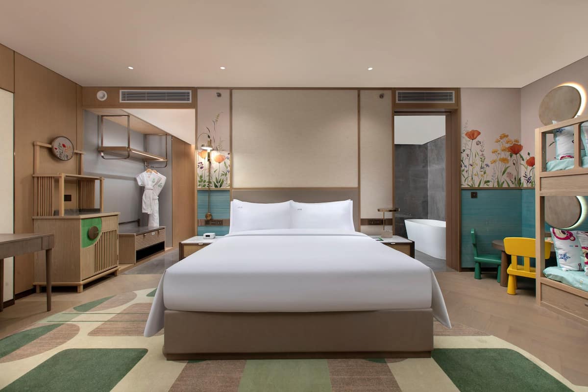 Una habitación de hotel moderna con una cama grande, un aparador de madera, un tocador con espejo, una mesita pequeña y una silla colorida.